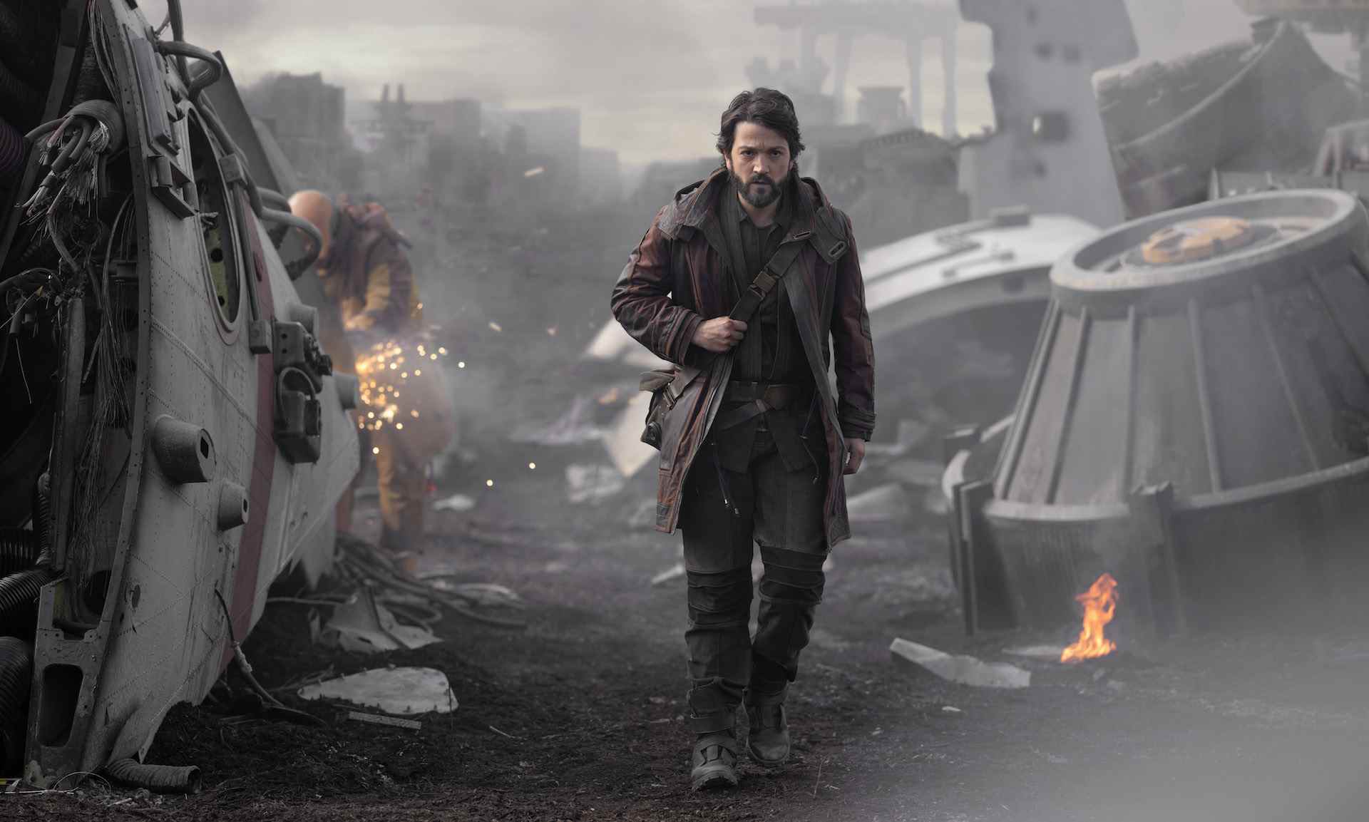 Un homme portant un manteau épais et plusieurs couches avec un sac sur l'épaule s'éloigne d'une scène de destruction;  encore de "Andor."