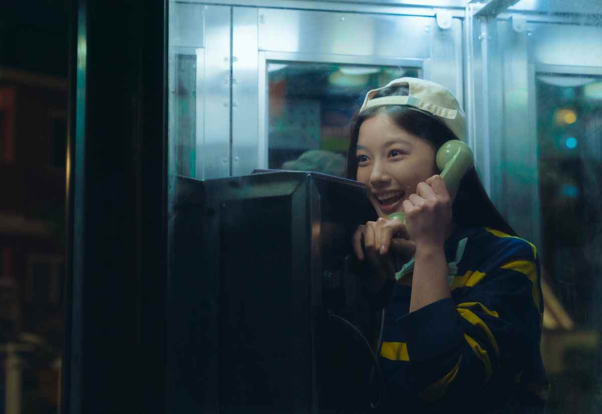 Une fille portant une casquette de baseball beige à l'envers sourit tout en parlant à la réception d'un téléphone vert à l'intérieur d'une cabine téléphonique en verre la nuit.