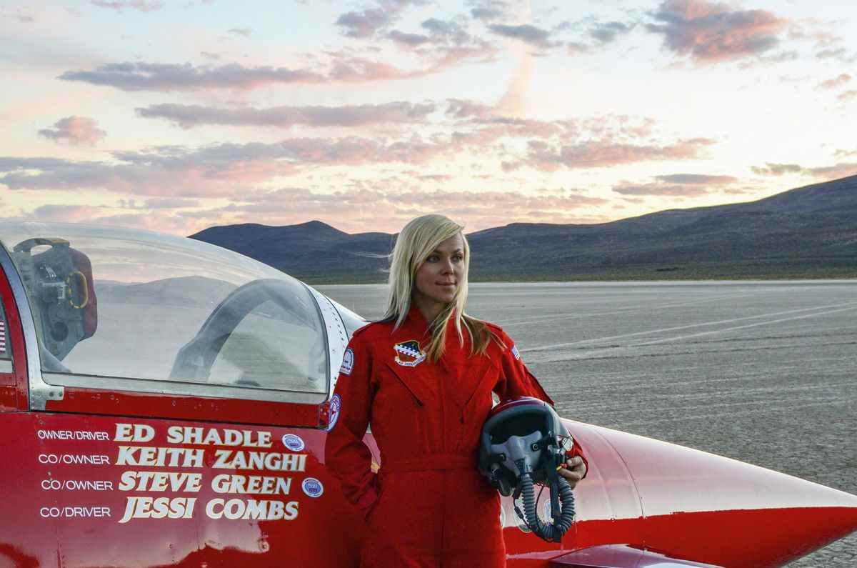Une femme aux cheveux blonds vêtue d'une combinaison rouge tient un casque de pilote debout devant le cockpit d'une voiture rouge au milieu d'un désert avec des nuages ​​ensoleillés au loin.