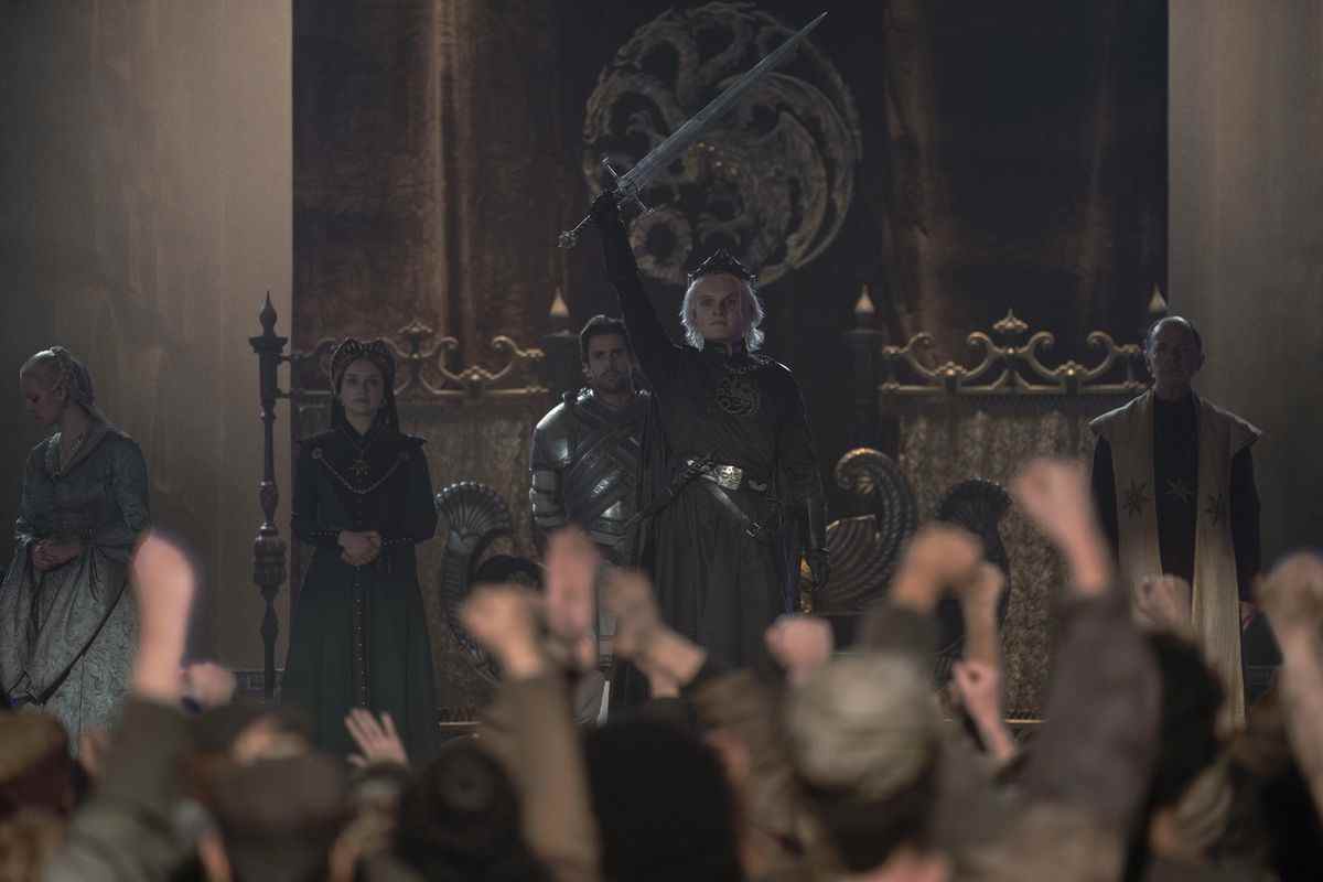 Le roi Aegon II (Tom Glynn-Carney) debout devant une foule de membres du public Westeros en liesse, tenant son épée en l'air triomphalement 