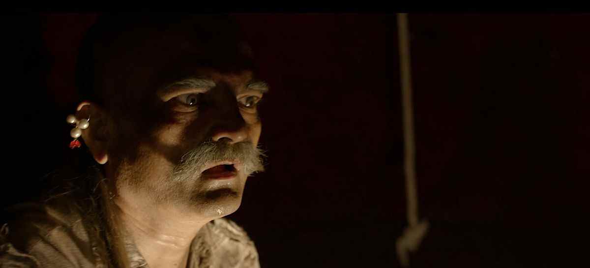 Un vieil homme avec une moustache de morse blanche et une boucle d'oreille en perles se tient dans le noir, bouche bée devant quelque chose hors écran, dans le film d'horreur indien Tumbbad