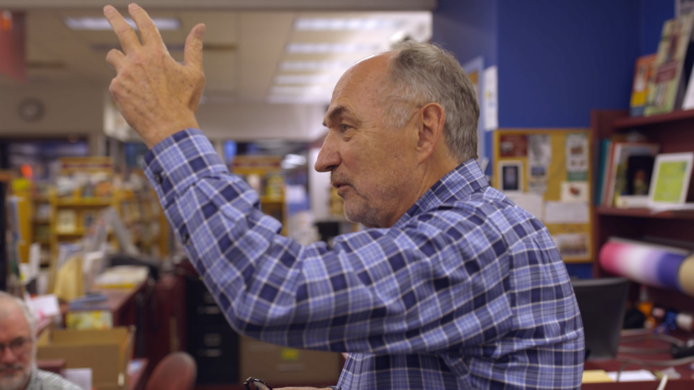 Un vieil homme en chemise bleue à carreaux lève la main et parle dans une librairie.