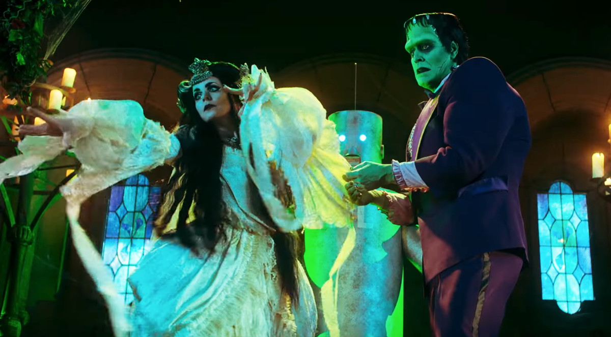 Une femme pâle aux longs cheveux noirs dans une robe blanche flottante se tient à côté d'un monstre Frankenstein à la peau verte dans un smoking violet.
