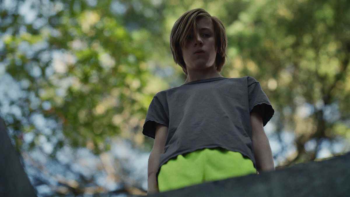John, 13 ans, avec des cheveux plus longs et un short jaune fluo, se tient au-dessus d'un trou avec la caméra le regardant dans John and the Hole