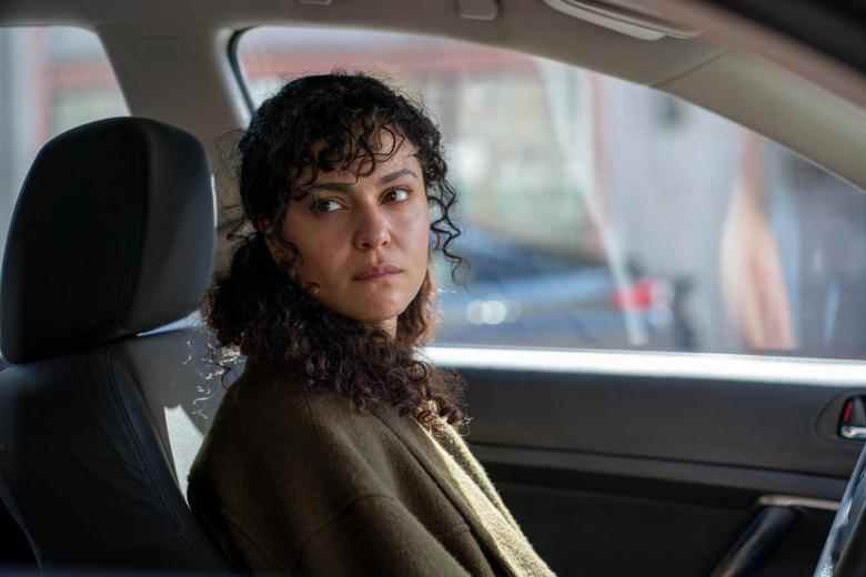 Une jeune femme aux cheveux bruns bouclés assise sur le siège du conducteur d'une voiture, regardant OS par la fenêtre du passager ;  encore de "Rami."