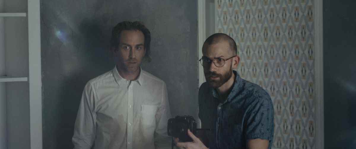Levi (Justin Benson) et John (Aaron Moorhead) se tiennent contre un mur aux motifs élaborés, pointant une caméra sur quelque chose hors écran qu'ils regardent avec méfiance dans Something In the Dirt