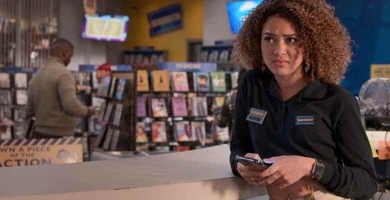 A teenage girl with curly brown hair in a navy blue Blockbuster Video shirt and khakis, standing holding her phone and looking disinterested in a Blockbuster video store; still from "Blockbuster."
