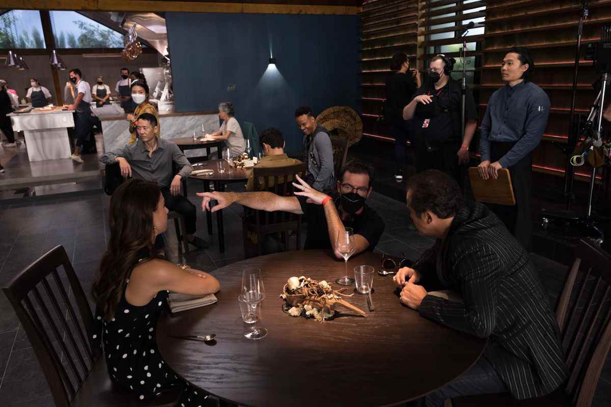Le réalisateur Mark Mylod est assis à une table sur le plateau avec plusieurs personnages de The Menu, faisant des gestes hors écran pendant qu'il dirige, avec l'équipement de caméra et les opérateurs en arrière-plan