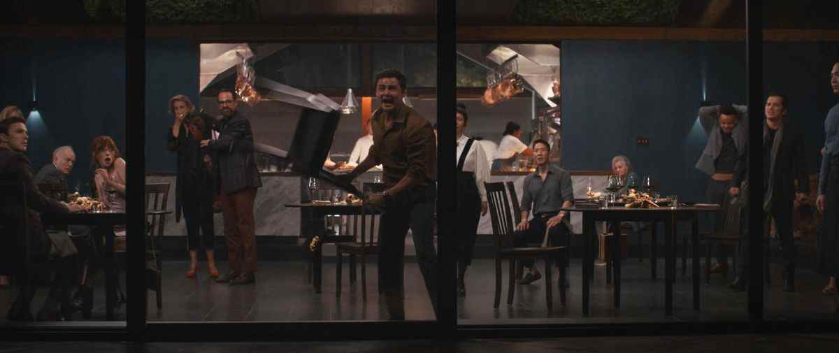 Un homme frénétique balance une chaise sur l'un des murs de verre d'un restaurant chic pour tenter de le briser et de s'échapper, tandis que le reste des clients effrayés regardent une scène de The Menu