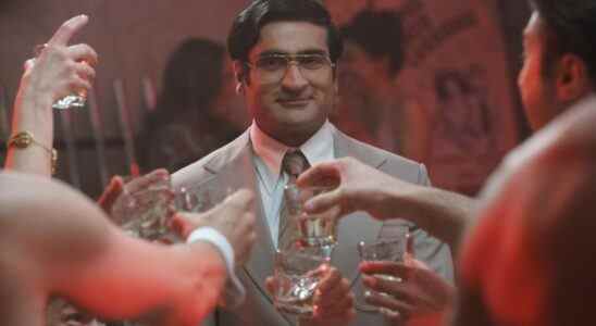 A man in a tan 1970s suit and large wire-rimmed glasses raising a glass along with others (only their hands in frame); still from "Welcome to Chippendales"