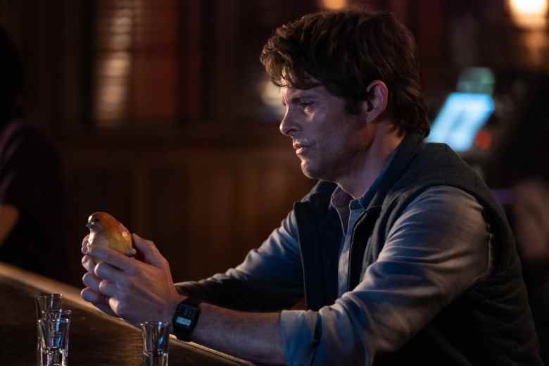 Un homme aux cheveux bruns ébouriffés assis dans un bar, portant une veste en cuir et regardant avec nostalgie un oiseau en bois cassé ;  encore de 