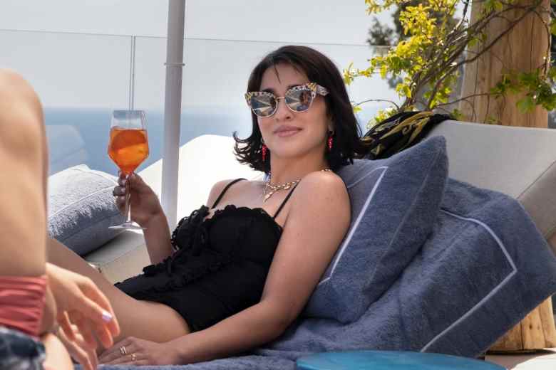 Une femme aux cheveux courts et foncés portant un maillot de bain une pièce noir et des lunettes de soleil, allongée sur une chaise de piscine et sirotant un aperol spritz au bord de l'océan ;  encore de 