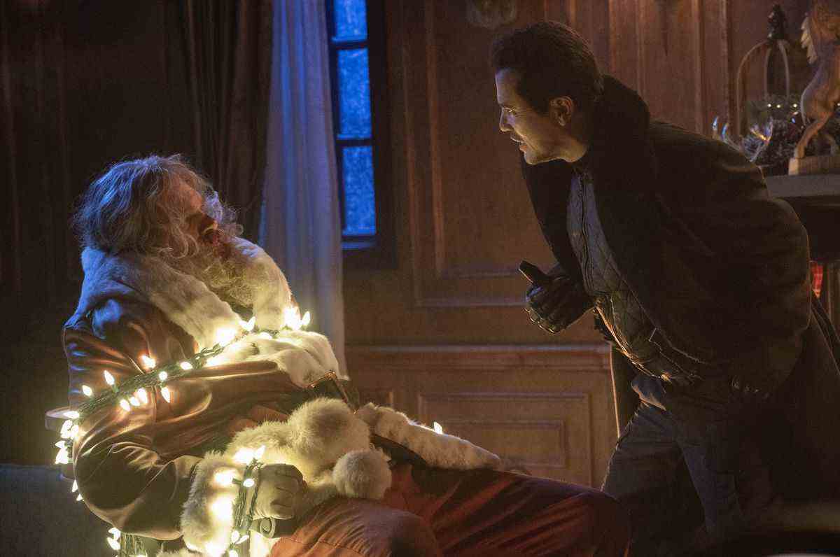 Scrooge (John Leguizamo) se penche d'un air menaçant sur le Père Noël (David Harbour), qui est attaché à une chaise avec une guirlande de lumières de Noël blanches allumées, dans Violent Night