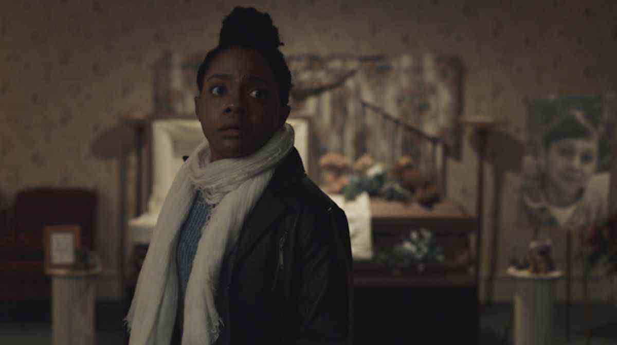 Monique (Gabby Beans), une femme à la peau foncée vêtue d'une veste noire et d'une écharpe blanche, se tient dans une pièce sombre avec un cercueil ouvert derrière elle et semble alarmée par quelque chose hors écran dans The Harbinger