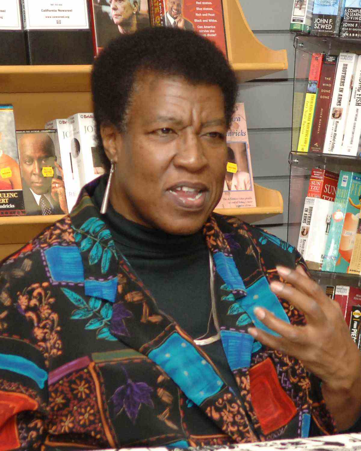Octavia E. Butler dans les années 1990, vêtue d'une chemise à motifs avec des motifs de feuilles d'arbres et de longues boucles d'oreilles en métal, est surprise au milieu d'une phrase lors d'une lecture dans une librairie