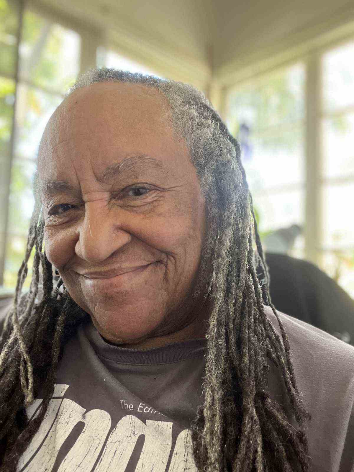 Andreea Kindryd, une femme noire plus âgée avec des dreadlocks grisonnants, sourit à la caméra avec des fenêtres très éclairées derrière elle