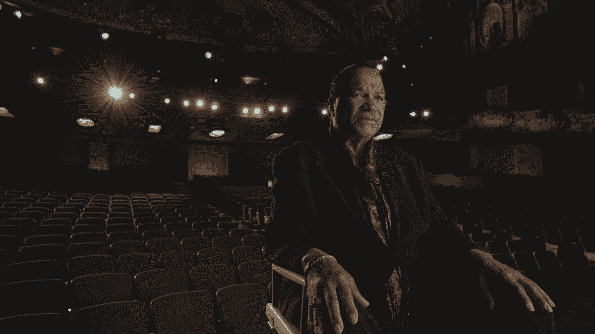 Billy Dee Williams est assis dans le fauteuil d'un metteur en scène sur une scène de théâtre, le dos face aux chaises vides du public