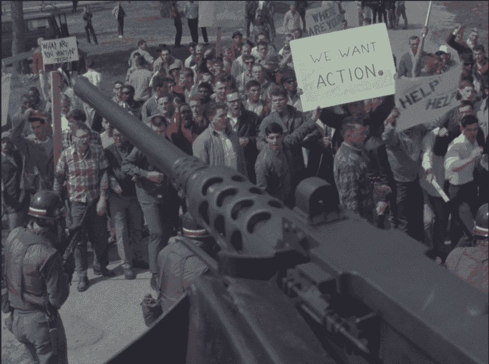 Un canon de char apparaît dans le cadre au-dessus d'une foule de manifestants mis en scène, dont l'un tient une pancarte indiquant 
