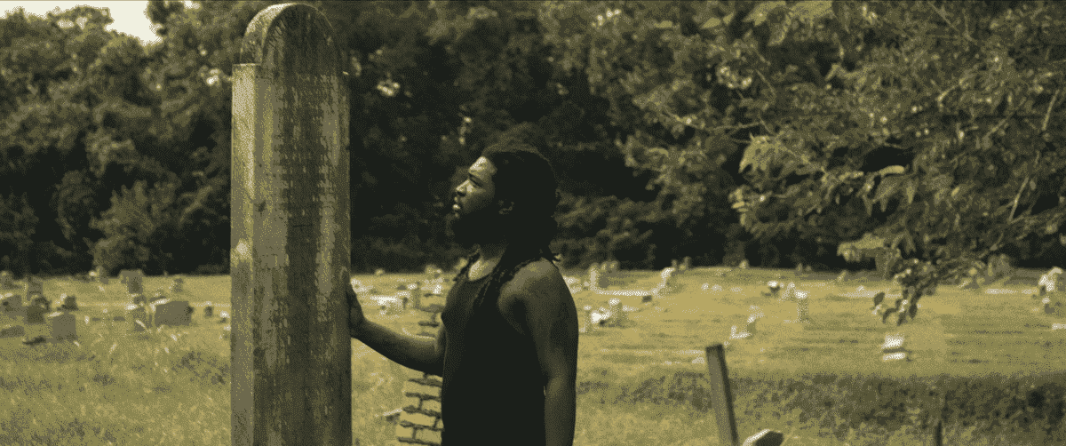 Un homme noir avec des dreadlocks et un débardeur noir se tient dans un cimetière en plaçant une main sur une imposante pierre tombale inscrite avec des noms