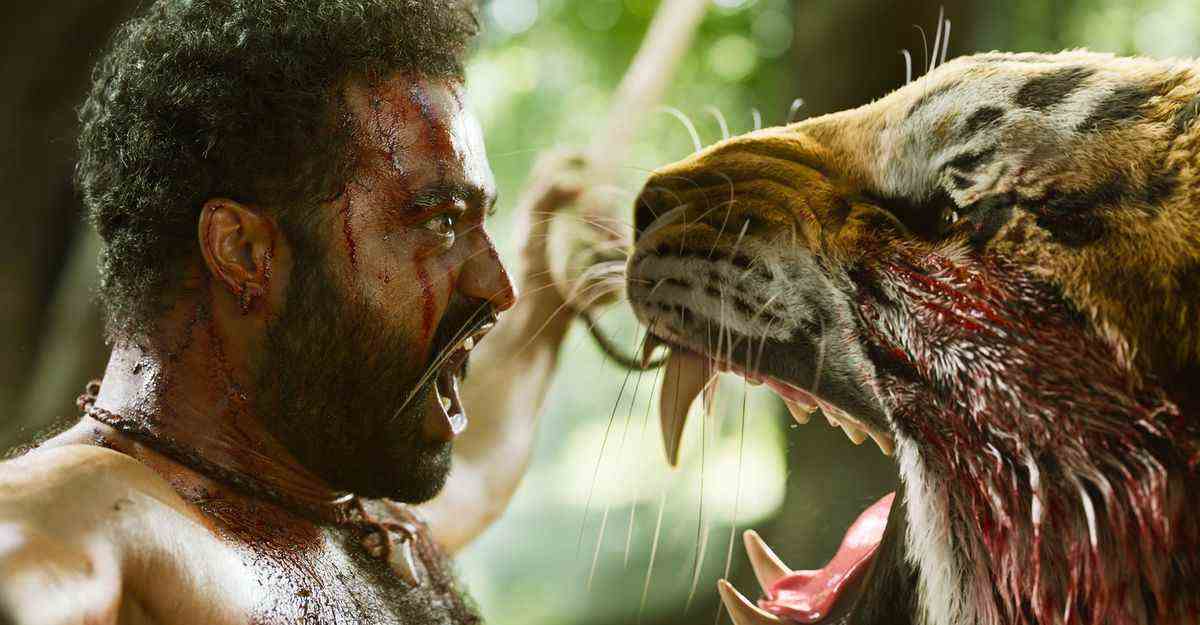 Bheem, un indien barbu, se tend en tenant les deux cordes d'un filet retenant un tigre, qui rugit au visage dans l'espoir de le mordre dans RRR