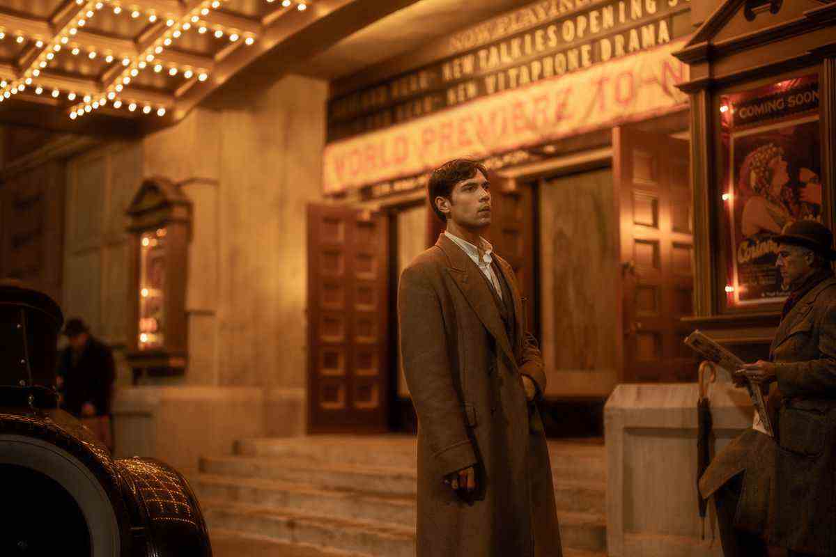 Manny se tient dans un trench-coat sous l'auvent d'un palais de cinéma, devant les affiches de chapiteau d'Hollywood classique dans le film Babylon