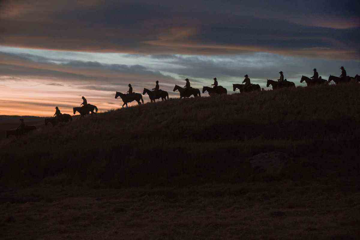 Une ligne de cow-boys chevauche leurs chevaux le long d'une crête contre le coucher du soleil dans une belle photo de la préquelle de Yellowstone 1923.