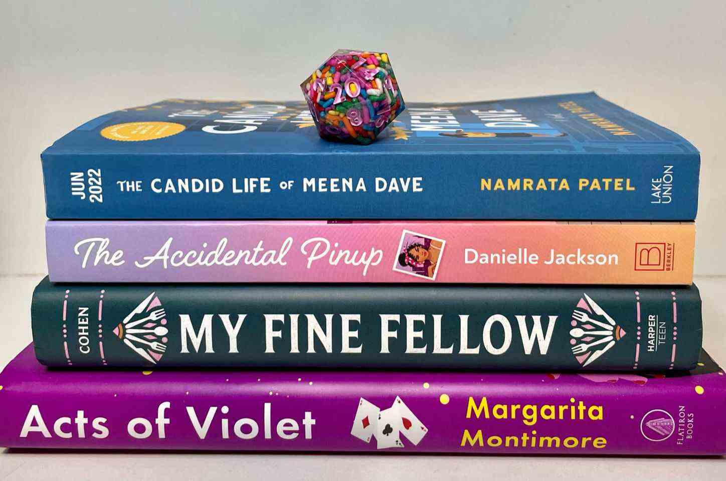 Un dé à 20 faces plein de paillettes avec des chiffres roses se trouve au sommet d'une pile de livres (The Candid Life of Meena Dave, The Accidental Pinup, My Fine Fellow et Acts of Violet)