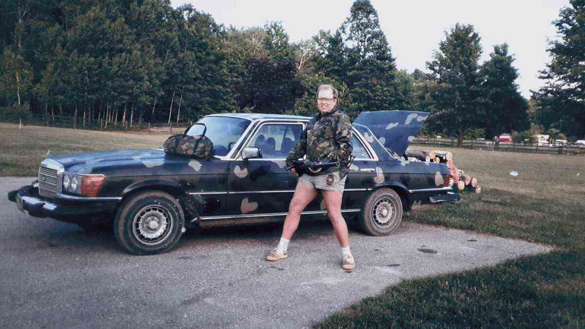 Une vieille photo des années 1980 montre un homme en camouflage et en short court traversant un champ jusqu'à un parking où sa berline camo est garée 