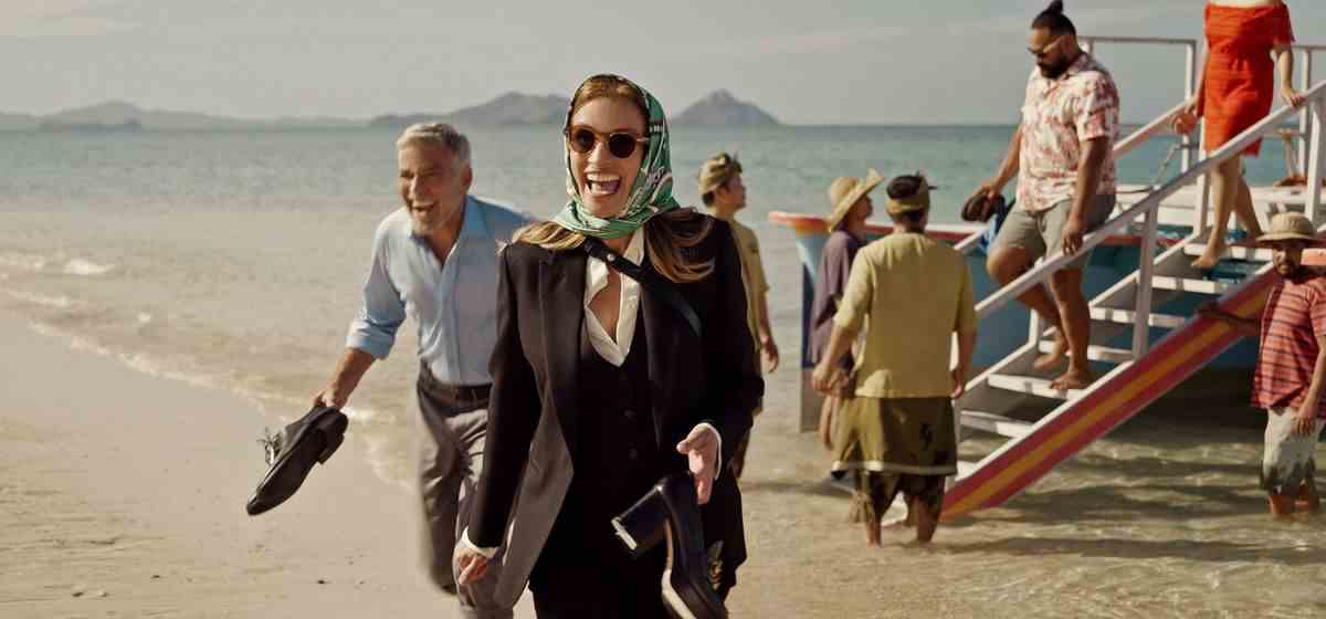 Un homme (George Clooney) et une femme portant un foulard sur la tête (Julia Roberts) rient en débarquant d'un bateau sur une plage et tenant leurs chaussures à la main.