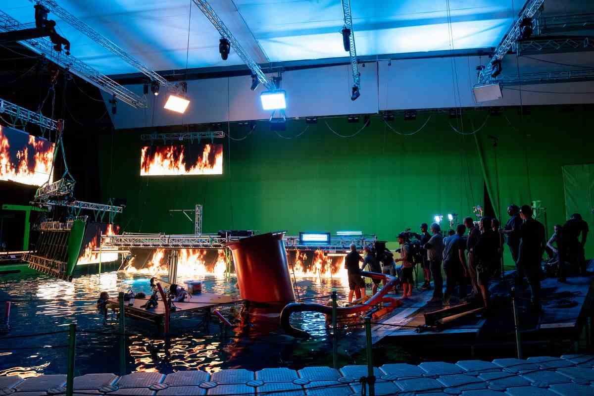 L'épave d'un navire construit sur une scène avec des écrans projetant des flammes photoréalistes et un fond d'écran vert.  James Cameron se tient sur une plate-forme avec son équipe dirigeant un acteur pendant le tournage d'Avatar : la voie de l'eau.