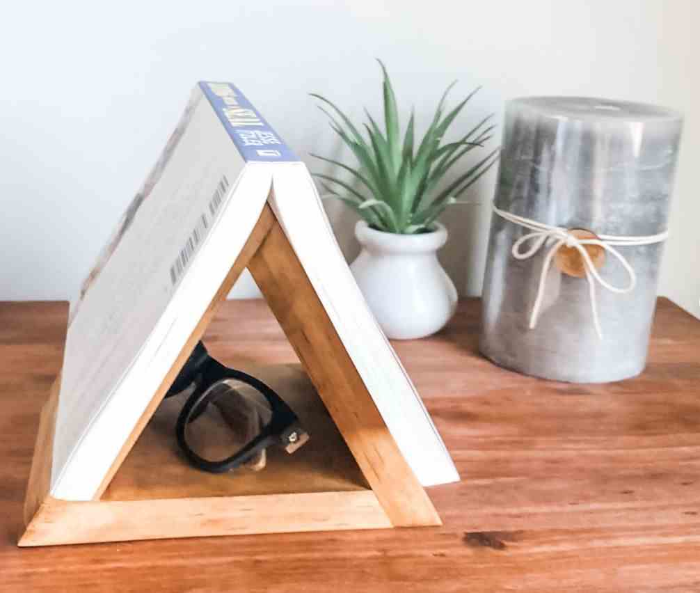 Image d'une table de chevet.  Sur celui-ci se trouve un triangle en bois qui permet à un livre de reposer ouvert sur le dessus, avec un espace pour les verres en dessous. 