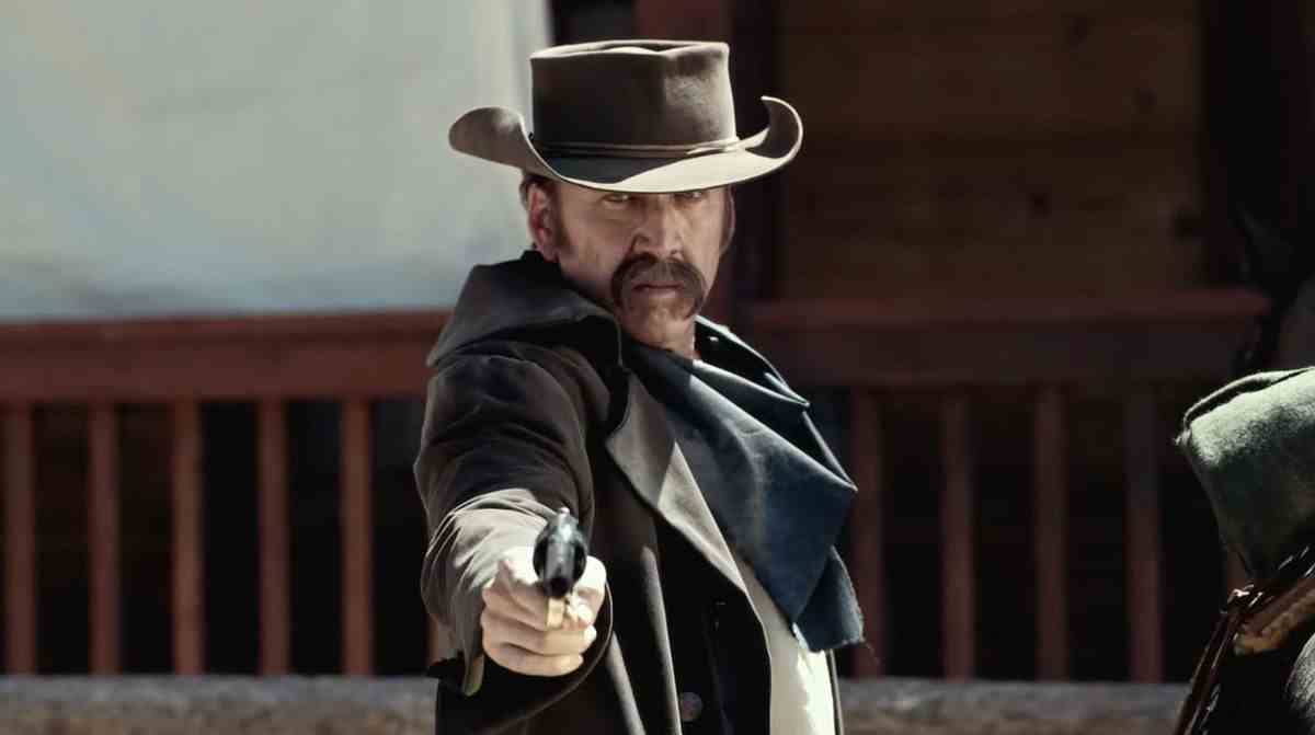 Un homme avec une moustache envahie (Nicolas Cage) portant un chapeau de cow-boy et un manteau de plumeau brun foncé visant un pistolet.