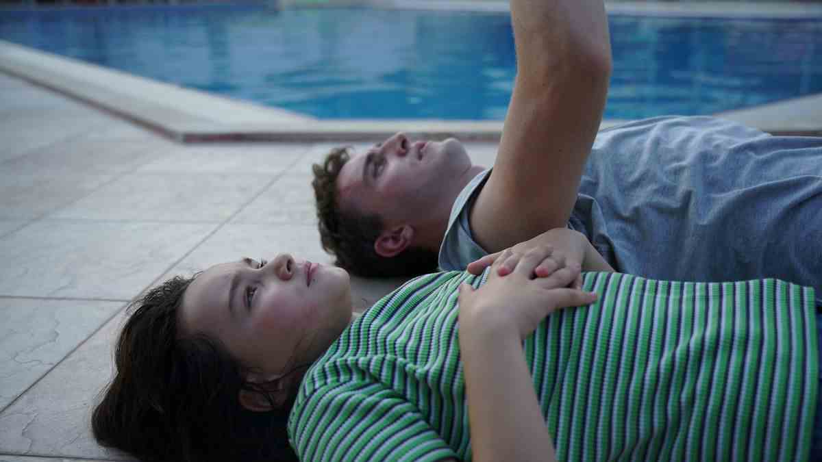 Un père et sa fille sont allongés sur le dos à côté d'une piscine tandis que le père montre le ciel dans Aftersun.