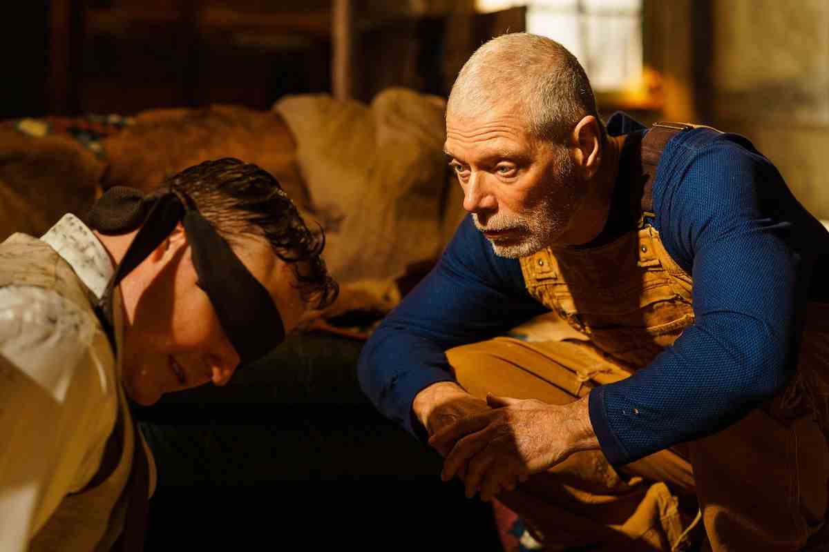 Un vieil homme (Stephen Lang) vêtu d'une chemise bleue à manches longues et d'une salopette jaune s'accroupit devant un homme aux yeux bandés (Marc Senter).