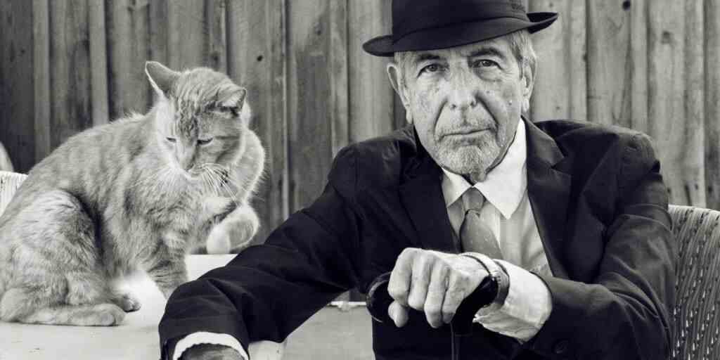 Une photo d'archive en noir et blanc de Leonard Cohen en costume sombre assis sur une chaise tenant une canne à côté d'une table avec un chat dessus.