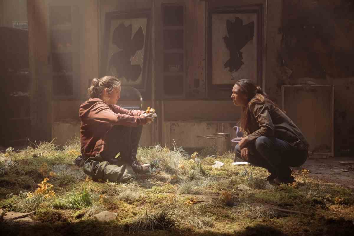 Ellie et Tess s'accroupissent face à face dans l'herbe qui a dépassé un bâtiment décrépit alors que la lumière brille d'en haut dans la série HBO The Last of Us.