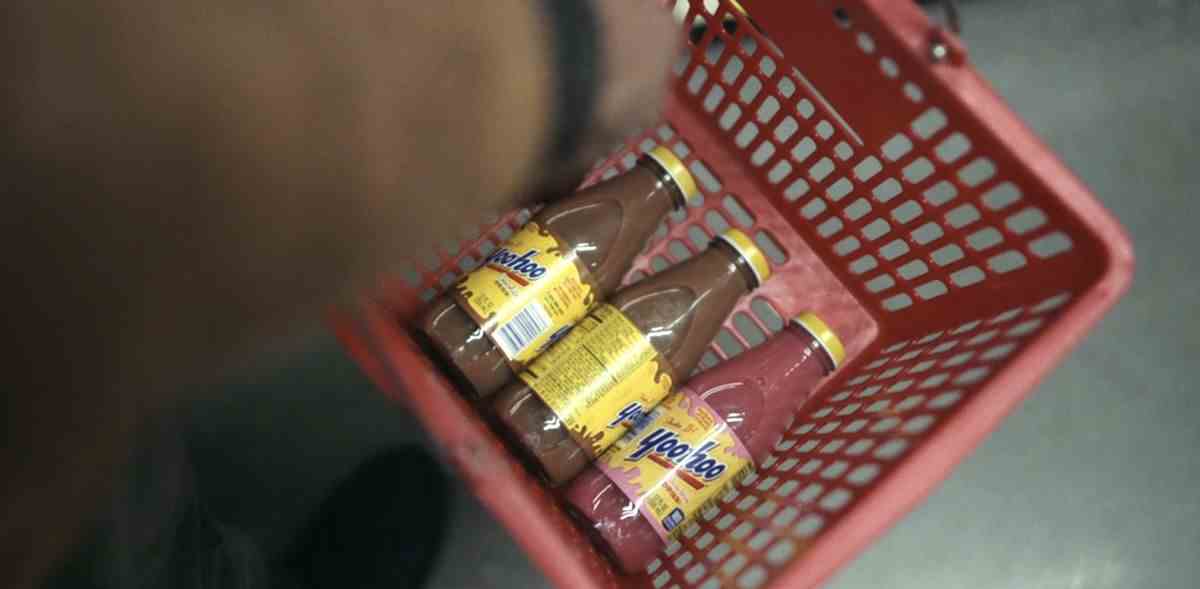 Un panier d'épicerie contenant deux bouteilles de chocolat Yoo-Hoo et une bouteille de fraise Yoo-hoo, tenu par un homme à la peau brune dans un manteau beige à Station Eleven