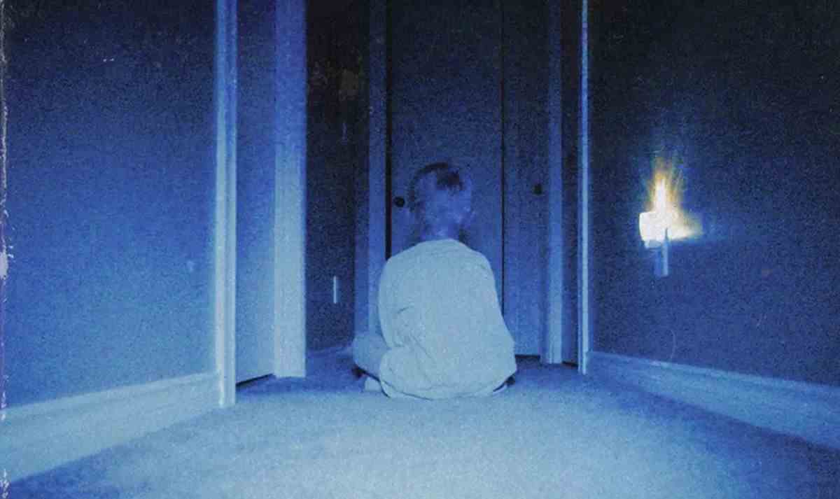 Un jeune garçon est assis dans un couloir bleu sombre, dos à la caméra, face à une série de portes ouvertes, dans un plan typiquement granuleux et flou du film d'horreur Skinarink