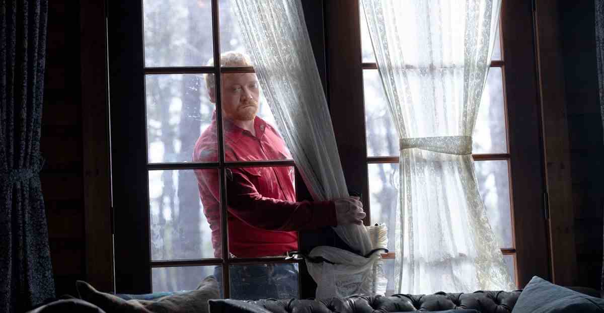 Redmond ( Rupert Grint ), un homme mince aux cheveux roux vêtu d'une chemise rouge vif, brise la vitre d'un ensemble de portes-fenêtres tout en forçant une entrée dans la cabine dans Knock at the Cabin