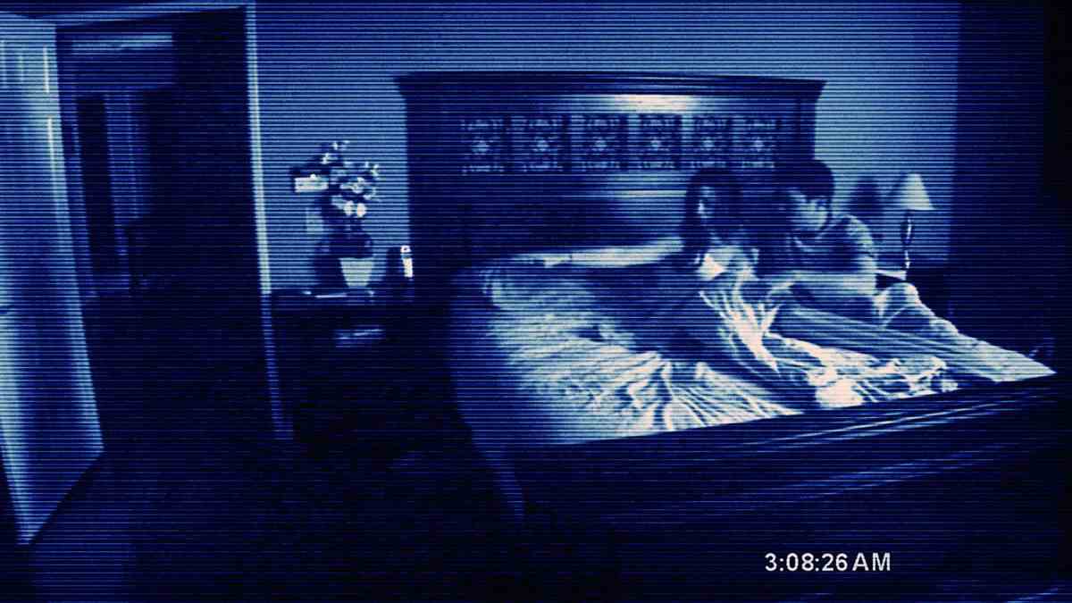 Un couple se blottit dans son lit, pointant avec crainte quelque chose hors écran, dans une image de caméra granuleuse et basse résolution de Paranormal Activity de 2007