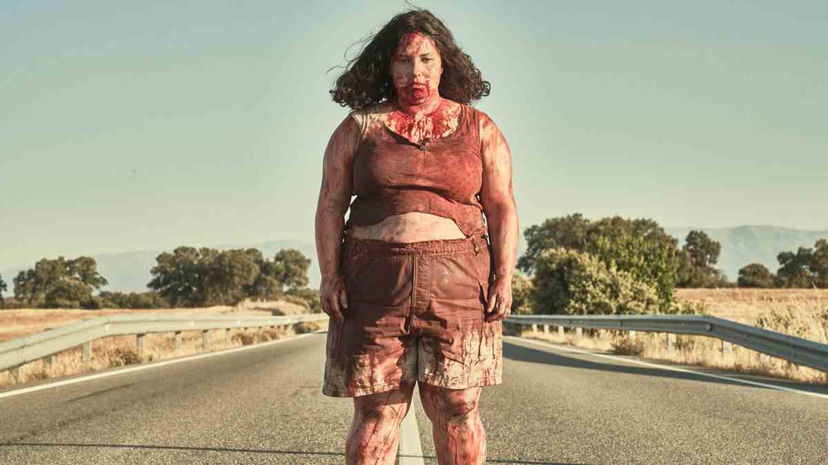 Une femme en maillot de corps blanc et en short se tient couverte de sang au milieu d'une route ouverte.