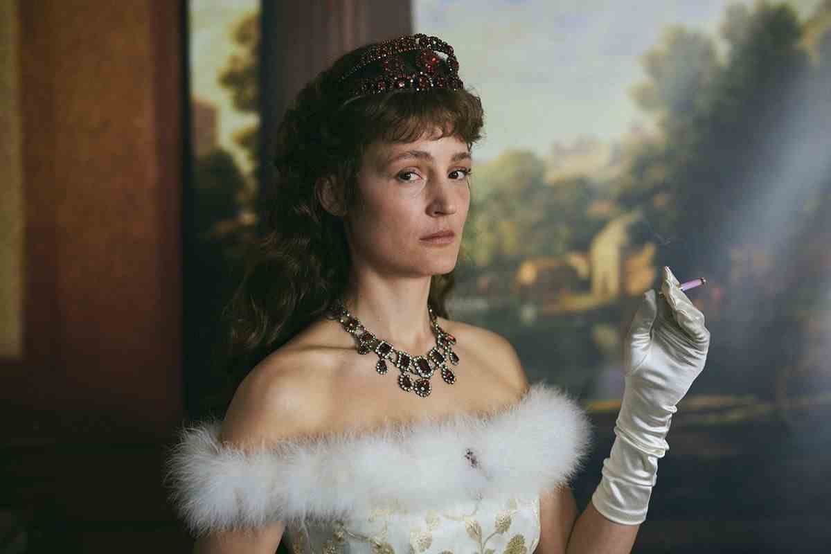 Vicky Krieps fume une cigarette et porte une couronne, une robe de fantaisie et un gant blanc devant une peinture ornée comme l'impératrice Elisabeth d'Autriche dans Corsage.