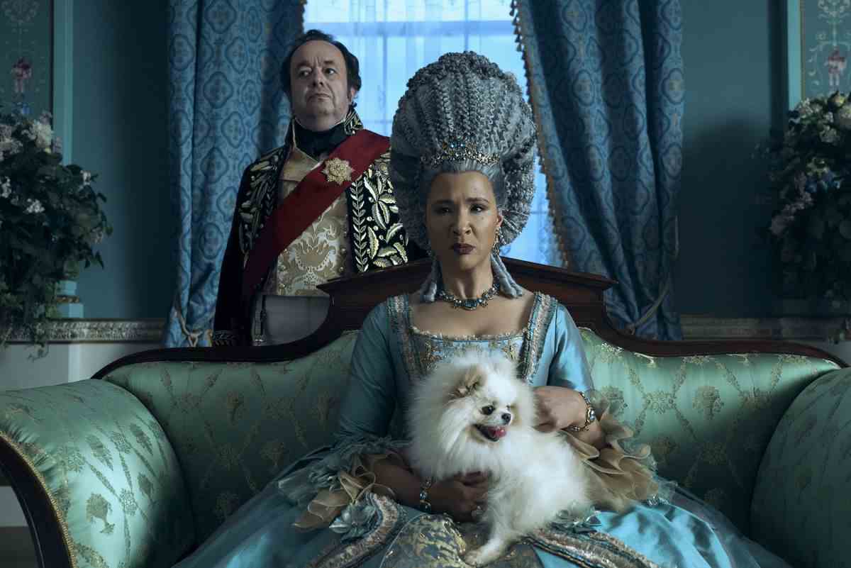 la reine charlotte plus âgée est assise sur un canapé chic.  les cheveux empilés, avec un chien blanc de fantaisie sur son dos.  derrière elle se trouve un fidèle intendant