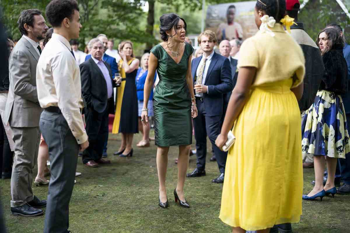 Une jeune femme noire vêtue d'une robe jaune vif se tient dans une foule de personnes à l'extérieur lors de ce qui semble être une garden-party et leur crie dessus dans une scène de The Strays de Netflix