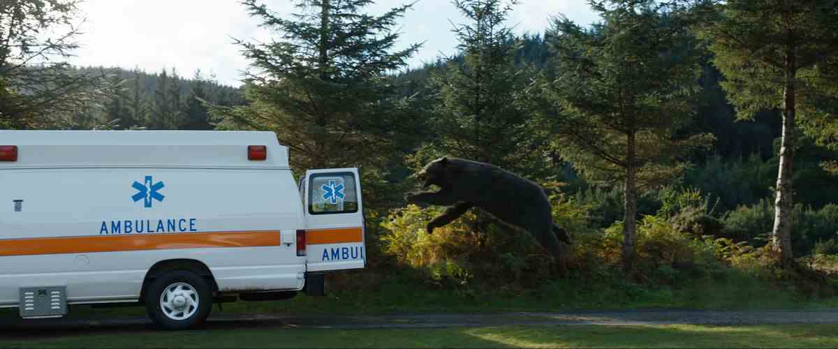 Un ours CGI fait un saut volant dans le dos ouvert d'une ambulance, vu dans un plan d'action à longue distance, dans Cocaine Bear