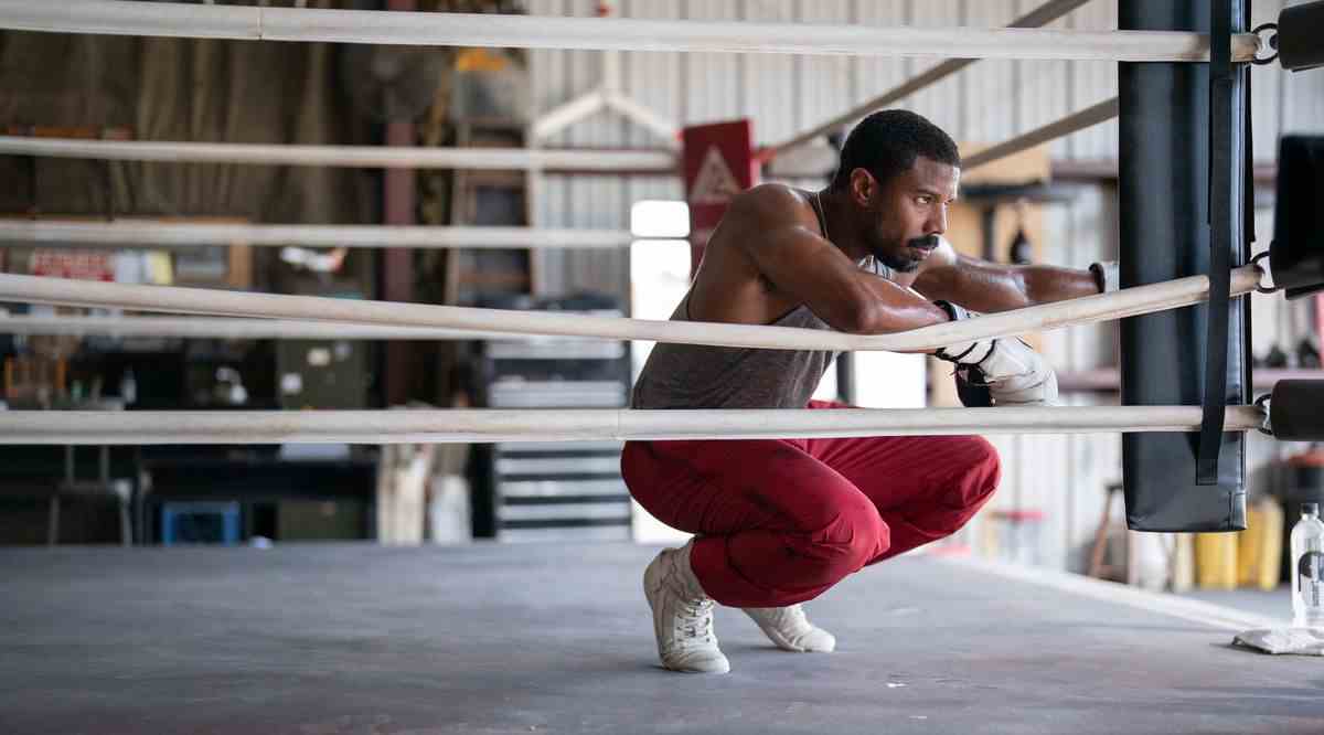 Adonis Creed (Michael B. Jordan), en survêtement et gants de boxe blancs, s'accroupit dans le coin d'un ring de boxe de gym et a l'air farouchement déterminé dans Creed III