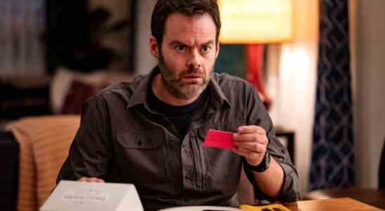 A man with dark hair and beard sits at a dining table, holding a red business card and looking horrified; still from "Barry."