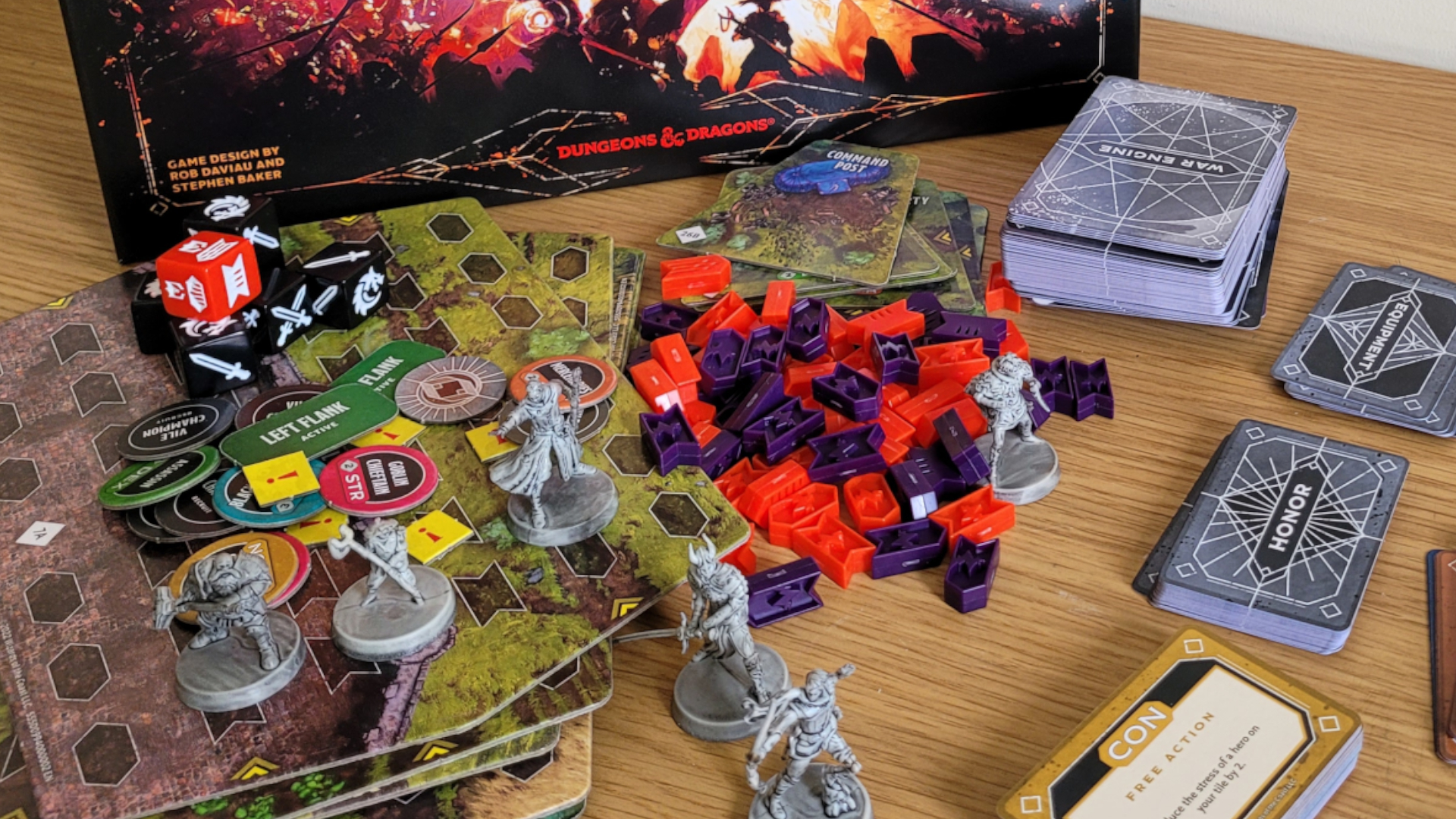 Les composants, les tuiles de plateau et les modèles de Dragonlance : Warriors of Krynn reposent sur une table en bois devant la boîte de jeu de société.