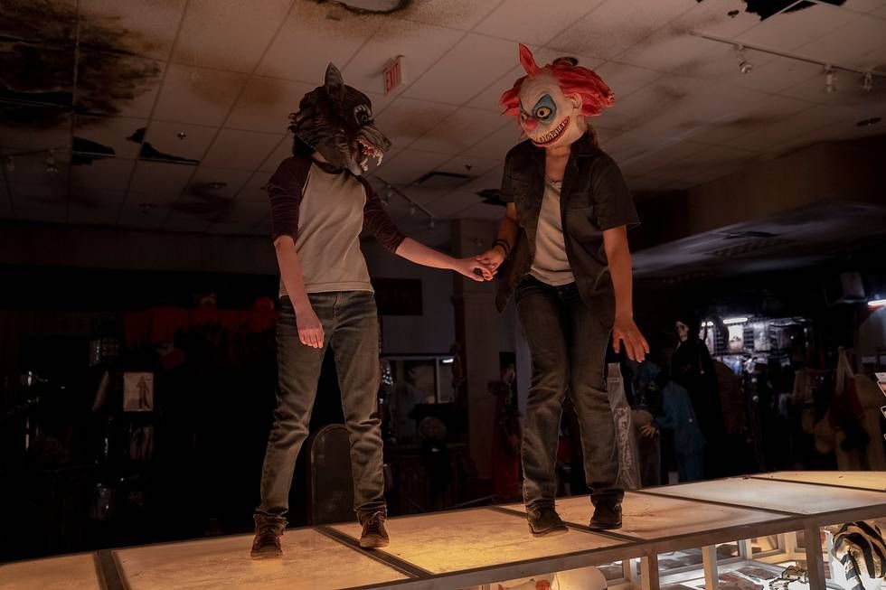 Ellie (Bella Ramsey) et Riley (Storm Reid) debout sur le comptoir d'un magasin d'Halloween et se tenant la main avec hésitation tout en portant respectivement un loup-garou et un masque de clown