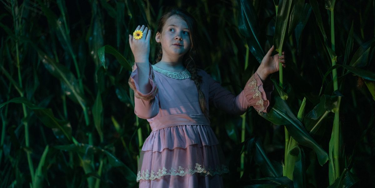 La méchante préadolescente et chef de secte Eden (Kate Moyer), une fille vêtue d'une robe rose sale avec de la dentelle au cou et aux poignets, se tient dehors dans un champ de maïs la nuit et lève une main arborant un anneau en plastique brillant en forme de tournesol dans le 2023 Enfants du maïs
